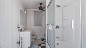 A compact bathroom with a square black and white checkered floor. It features a glass-enclosed shower, a sink with a mirror above, a toilet, a ceiling fan, and a small window with blinds. Some toiletries are visible on the sink.