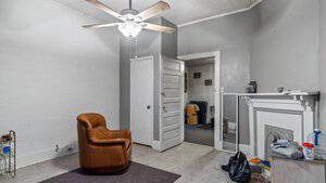 A room with a brown recliner, ceiling fan, white walls, and gray accent wall. There's a small table in the corner, a closed door, and an open door leading to another room with a bed. A fireplace is on the right with a blue vase and some items on it.
