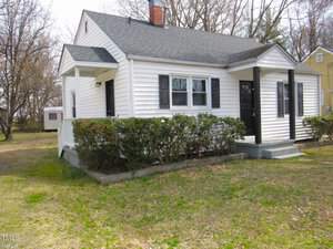 A small white house with a gabled roof and black trim is surrounded by a yard and bushes. There is a narrow porch with a black door, and a chimney on the roof. Leafless trees are in the background.
