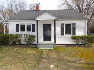 Nestled at 734 Baker Street in Oxford, this small, white, single-story house boasts a black door and shutters. With a shingled roof and chimney, it features a quaint front stoop with steps leading to a path. Shrubs border the yard, while trees provide a lush backdrop.