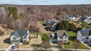 Aerial view of a suburban neighborhood featuring several houses with driveways and yards. Some homes have fences and decks. The background shows a wooded area with bare trees under a partly cloudy sky.