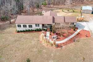 Aerial view of a single-story house with a brown roof, surrounded by a landscaped garden with red mulch, shrubs, and a paved walkway. The yard is expansive with dry grass, and a small structure is visible in the backyard near the trees.