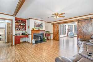 Living room with wood flooring, a ceiling fan, and a fireplace. There are barn-style sliding doors leading to a sunlit room with large windows. A red built-in shelf with decorative items is next to the fireplace, and there's a leather couch.