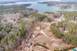 Aerial view of a rural area featuring a large house with a red roof surrounded by trees. A winding driveway leads to the house. There's a nearby body of water in the background and several smaller structures scattered around the property.