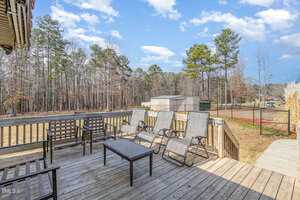 Wooden deck with six reclined lounge chairs, two black benches, and a central table. The deck overlooks a grassy yard with surrounding trees under a partly cloudy sky. In the background, there are storage sheds and a fenced area.