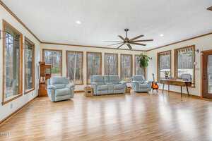A spacious sunroom with large windows overlooking a forest. The room features two armchairs and a sofa in light fabric, a ceiling fan, a small table with a chessboard, a guitar, a potted plant, and wooden flooring.