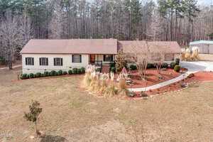 A single-story house at 89 Mount Pleasant Lane has a beige exterior and brown roof, nestled in a yard with minimal grass. The property features a paved walkway, small shrubs, and mulch landscaping, surrounded by tall trees stripped of leaves.