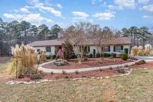 A single-story light-colored house with a brown roof surrounded by landscaped gardens. The yard features ornamental grasses, a stone path, a small fountain, and a picnic table with a red umbrella. Tall trees line the background under a partly cloudy sky.