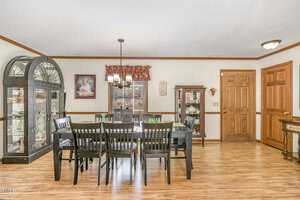 A dining room with a wooden table and eight matching chairs. On the left, a large cabinet with glass doors displays various items. A smaller cabinet is to the right. A window with a curtain sits in the middle. The floor is light wood.