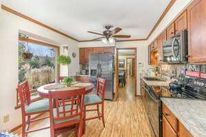 A kitchen with wood cabinets, stainless steel appliances, and a round dining table with four chairs. The room has hardwood floors, a ceiling fan, a bay window with plants, and access to a dining area in the background.