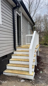 A small wooden staircase with white railings leads to the side entrance of a gray building. The door is open, revealing a glimpse of the interior. The surrounding area has bare ground and scattered debris, with trees in the background.