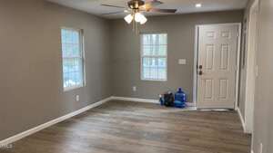 Empty room with light gray walls and wooden flooring. Features a ceiling fan with lights, two windows, and a closed white door. A filled blue water jug and some bags are placed near the door next to an electrical outlet.