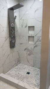 Marble-tiled shower with hexagonal floor tiles. A vertical shower panel with multiple controls and a handheld showerhead is mounted on the wall. There are two recessed shelves in the wall.