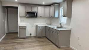 A modern kitchen with light gray cabinets, a marble countertop, and a stainless steel sink. There is a built-in microwave above the counter. The floor is wood, and there is a window above the sink letting in natural light.