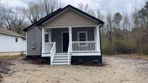 A small, single-story house with gray siding and a porch featuring white railing stands quietly at 471 Merriman Street. The yard is bare dirt with a few plants by the foundation. In the background, trees reach for cloudy skies in charming Henderson.