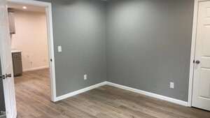 A small, empty room with gray walls and light wood flooring. There is a door to the left leading to another room with white walls. A closed white door is on the right. Several electrical outlets are visible on the walls.
