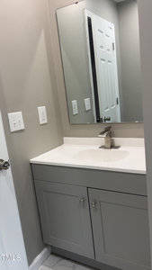 A bathroom with a white countertop and gray vanity cabinet. A rectangular mirror is mounted above the sink. The wall is painted light gray, and there are two switches on the left wall. Part of a white door is visible.