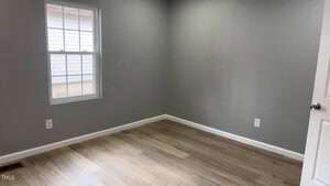 An empty room with gray walls and light wood flooring. There is a single window on the left wall, showing the exterior siding of another building. A white door is partially visible on the right side. The room has white baseboards and electrical outlets.
