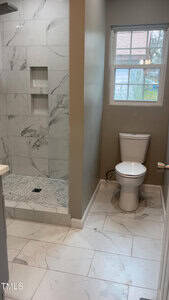 Bathroom with a tiled walk-in shower featuring built-in shelves on the left. A white toilet is positioned under a window with a view of greenery. The floor is covered with large white tiles, and the wall paint is beige.
