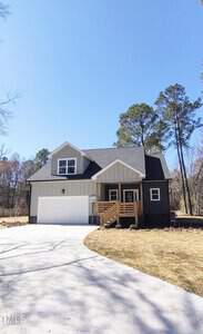 Located at 307 7th Street, this charming two-story house in Butner boasts a gray roof and beige siding. A white garage door complements the wooden front porch, all set against a backdrop of trees under a clear blue sky. The concrete driveway elegantly leads to the garage.