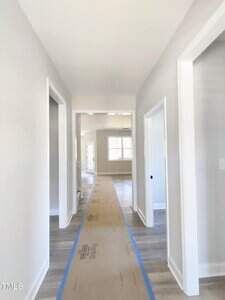 A hallway in a new house with light-colored walls and wooden flooring. A protective cardboard strip runs along the floor, leading to a bright room with large windows in the background. Three doorways are visible on the sides.