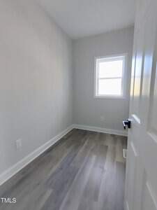 Small empty room with light gray walls, a wood-grain laminate floor, and a white trim window. The room is seen from an open white door with a black handle. No furniture is present.