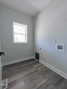 Small, empty room with light gray walls and vinyl plank flooring. A window on the left wall lets in natural light. There's a plumbing and electrical setup for a washer and dryer on the right wall.
