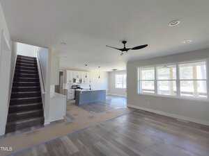 A modern, unfurnished open-plan living space featuring a kitchen with an island, stainless steel appliances, and pendant lights. The room has wooden floors, large windows, a ceiling fan, and a view of a staircase leading to the upper floor.