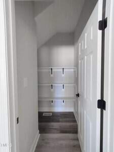 A small, empty walk-in closet with white walls and ceiling, featuring wooden flooring. Three white shelves with dark brackets are mounted on the back wall. The closet door is open, revealing a white frame and black hinges.