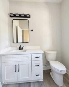 A bathroom with white walls features a rectangular mirror and a modern light fixture above it. A white vanity with black handles holds a sink and faucet. Next to it is a toilet. The floor is light-colored wood or laminate.