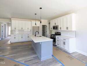 A modern kitchen with white cabinetry, a central island, and stainless steel appliances. The floor is light wood, and the countertops are white. Three black pendant lights hang above the island, and cardboard is laid out on the floors.