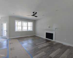 A vacant living room with light gray walls and wood laminate flooring. It features a black ceiling fan, a white fireplace with a mantel, and a large window. The floor is partially covered with protective paper near the entrance.