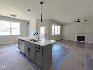Modern open-concept kitchen and living room. The kitchen features a large island with a sink and pendant lights. The living area has large windows and a fireplace. Floors are covered for protection. Walls are painted light gray.