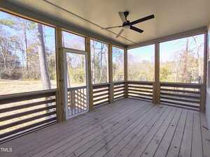 A screened porch with wooden flooring and railing, featuring a white ceiling with a black ceiling fan. The porch has large screened windows and a screen door, offering views of a grassy yard and trees.