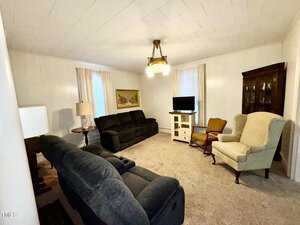 A living room with white walls and two dark sofas facing each other. A TV sits on a white stand. There are two armchairs and a small table. A chandelier hangs from the ceiling, and a framed picture is on the wall. Curtains cover two windows.