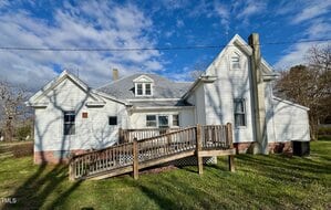 A white, single-story house with a gabled roof and multiple extensions is shown against a blue sky with clouds. The house has a wooden ramp leading to the front entrance and is surrounded by a grassy yard. A wooden utility pole is in the foreground.
