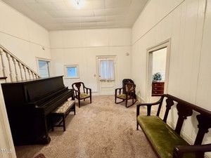 A small, brightly lit room with white paneled walls and carpeted floor. It contains a black upright piano, two wooden chairs with green velvet cushions, a matching bench, and a wooden armchair. There are three doorways and a staircase is partially visible.