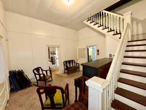 A room with white walls features several wooden chairs and a small wooden cabinet. A staircase with white railings leads upward. Folds of chairs are stacked in one corner, and a doorway opens to another room. Carpet covers the floor.
