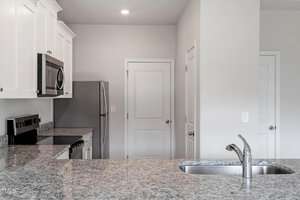Modern kitchen with stainless steel appliances, including a refrigerator, oven, and microwave. White cabinetry contrasts with gray granite countertops. A double-basin sink and faucet are in the foreground. Two closed doors in the background.