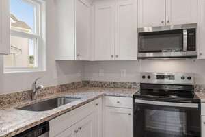 A modern kitchen with white cabinets and granite countertops. On the right, there is a stainless steel stove and microwave. A single sink with a chrome faucet is under a window on the left.
