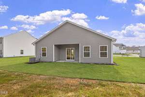 A single-story gray house features a gable roof and a small covered patio with double glass doors. Two windows flank the doors, and the house is surrounded by a grassy yard. Neighboring houses are visible on both sides under a partly cloudy sky.