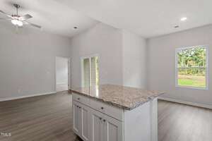 Modern open-concept living area with neutral walls and wood flooring. A kitchen island with a granite countertop is prominent in the foreground. A ceiling fan hangs from the recessed ceiling. A window offers a view of a grassy landscape.