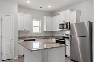 A modern kitchen with white cabinetry and stainless steel appliances, including a refrigerator and microwave. An island with a granite countertop is central. A sink below a small window and a closed pantry door are also visible.