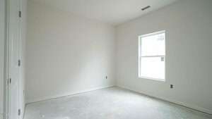 An empty room with white walls and a concrete floor. There is a single window on the right wall allowing natural light in. The room appears unfinished, with visible outlets and no furnishings.