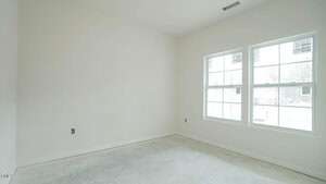 An empty, unfurnished room with unpainted walls and a concrete floor. Two large windows on the right side let in natural light. The space is under construction or renovation, with outlets and unfinished details visible.