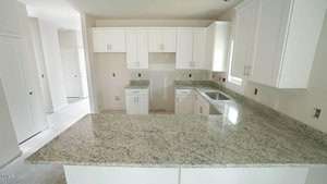 A modern kitchen with white cabinets and light gray speckled granite countertops. There is a stainless steel sink under a window. The kitchen has no appliances installed. Walls are painted white. A hallway and open door are visible to the left.