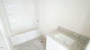Bathroom under construction with unfinished gray concrete floor. Includes an alcove bathtub with three built-in shelves on each side and a white vanity with a granite countertop and white undermount sink. Walls are painted white.