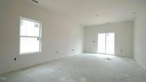 An empty room with unfinished floors, white walls, and a window on the left. There is a sliding glass door on the right leading outside, allowing natural light. The room is bare, with visible electrical outlets and ventilation openings.