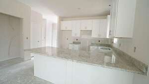 A modern kitchen with light-colored granite countertops, white cabinetry, and unfinished flooring. A window above the sink provides natural light. Walls are unpainted with visible outlets and light fixture placements.