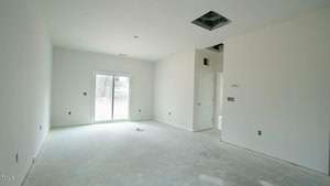 Empty room with unpainted walls and concrete floor. A sliding glass door on the left opens to the outside. A small square ceiling cutout is visible. An open doorway leads to another room on the right. Sparse light fixtures and outlets are installed.